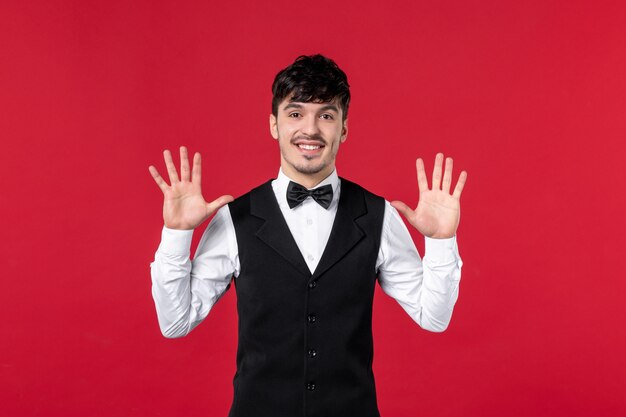 Front view of of smiling male waiter in a uniform with bowtie on neck and showing ten on red wall