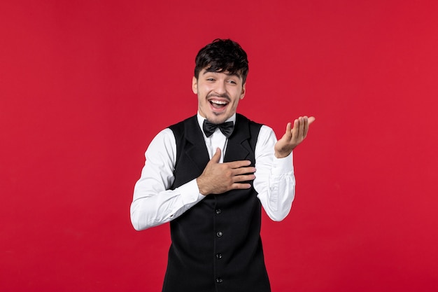 Front view of smiling male waiter in a uniform with butterfly on neck and feeling curious about something on red background