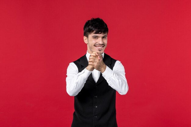 Front view of smiling male waiter in a uniform with butterfly on neck and feeling happy about something on red background