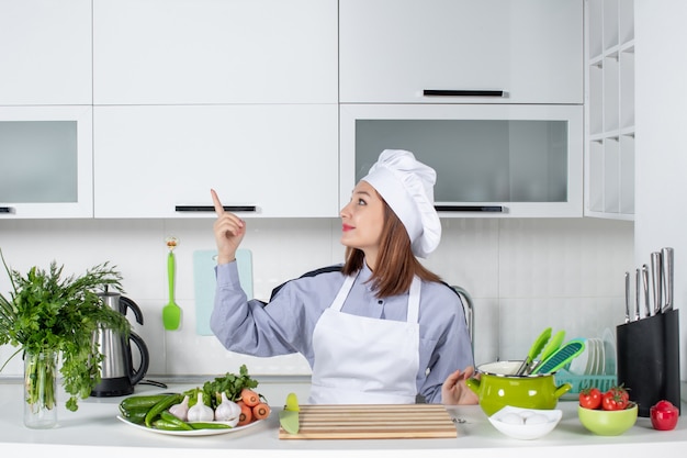 Front view of smiling positive female chef and fresh vegetables pointing up on the right side in the white kitchen
