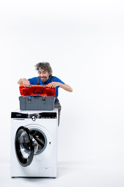 Front view of smiling repairman opening tools bag behind washer on white wall