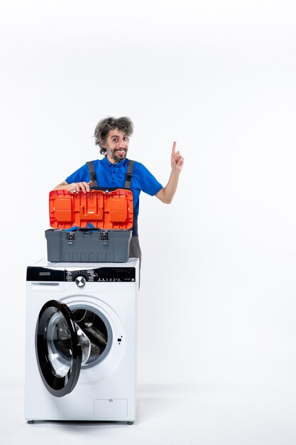 Front view of smiling repairman opening tools bag behind washing machine on white isolated wall