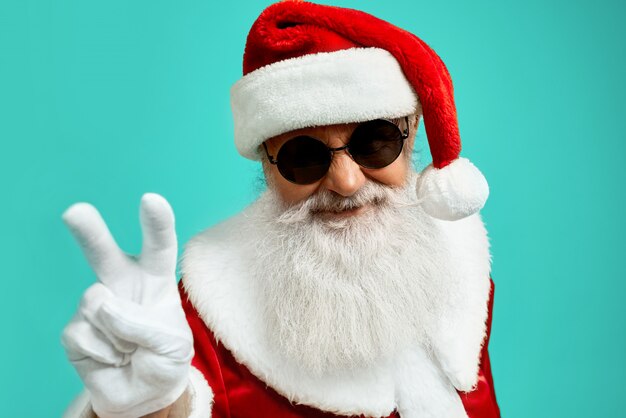 Front view of smiling Santa Claus with long white beard showing peace with two fingers up. Funny senior stylish man in sunglasses posing 