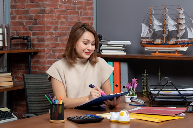 Front view smiling woman using highlighter sitting at wall