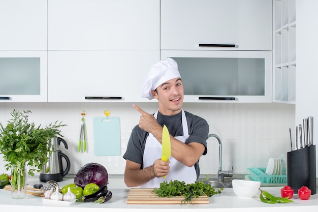 Front view smiling young chef in uniform standing behind kitchen table