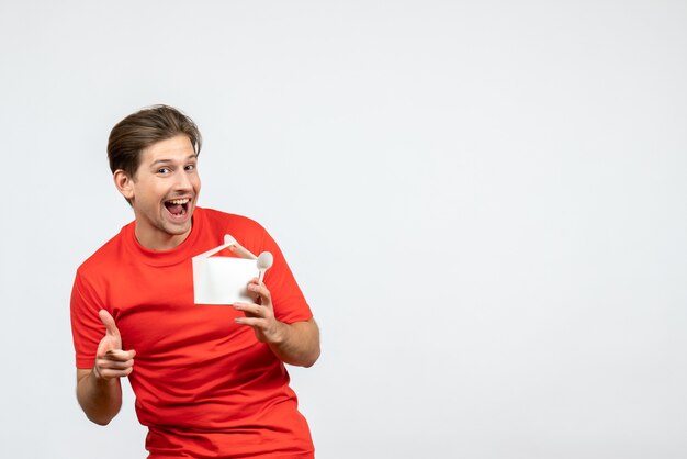 Front view of smiling young guy in red blouse holding paper box and spoon pointing forward on white background