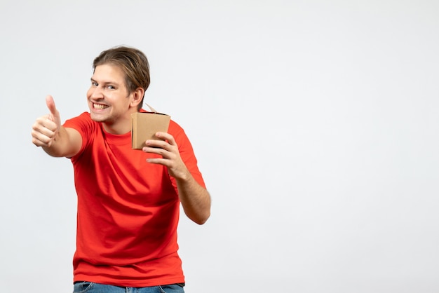 Front view of smiling young guy in red blouse holding small box making ok gesture on white background