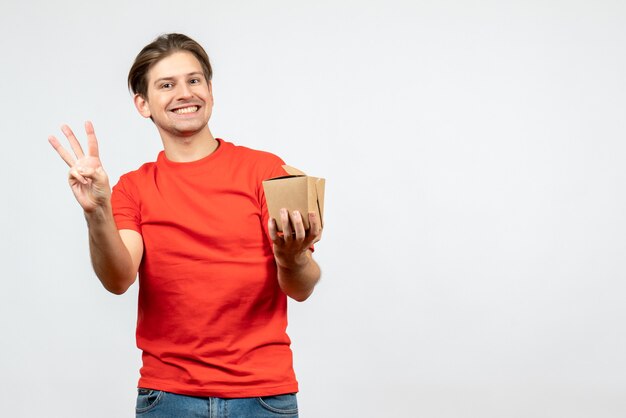 Front view of smiling young guy in red blouse holding small box showing three on white background