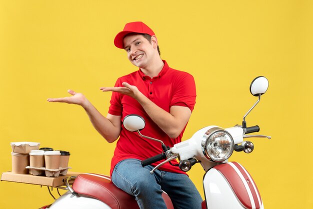 Front view of smiling young guy wearing red blouse and hat delivering orders pointing something on the right side on yellow background