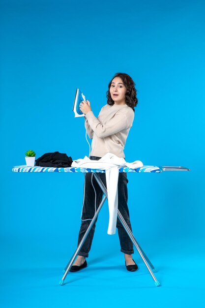 Front view of smiling young lady standing behind the board and ironing the clothes on blue background