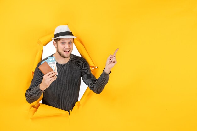 Front view of smiling young man with a hat holding foreign passport with ticket and pointing up in a torn on yellow wall