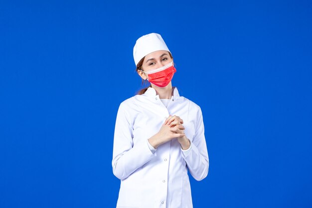 Front view smiling young nurse in medical suit with red protective mask on blue 