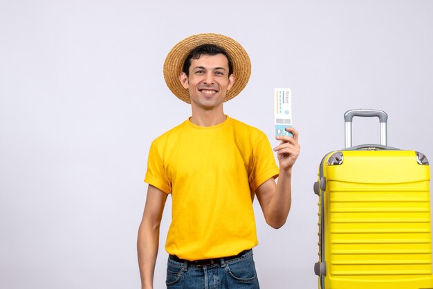 Front view smiling young tourist standing near yellow suitcase holding ticket