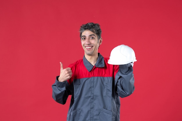 Foto gratuita vista frontale del giovane lavoratore sorridente in uniforme e tenendo il casco e facendo un gesto ok sul muro rosso isolato