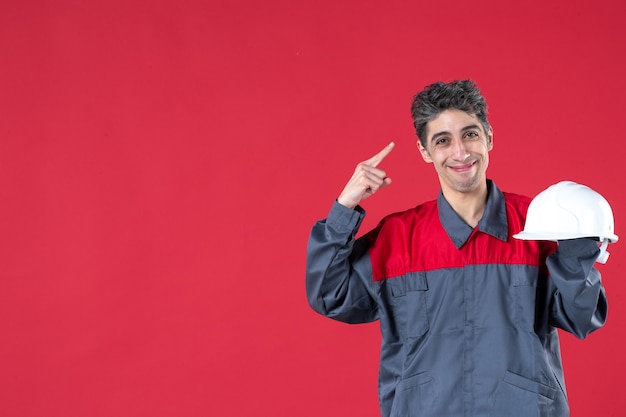 Front view of smiling young worker in uniform holding hard hat and pointing himself on isolated red wall