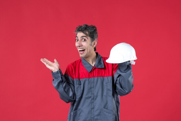 Front view of smiling young worker in uniform and holding hard hat pointing something on the right side on isolated red wall