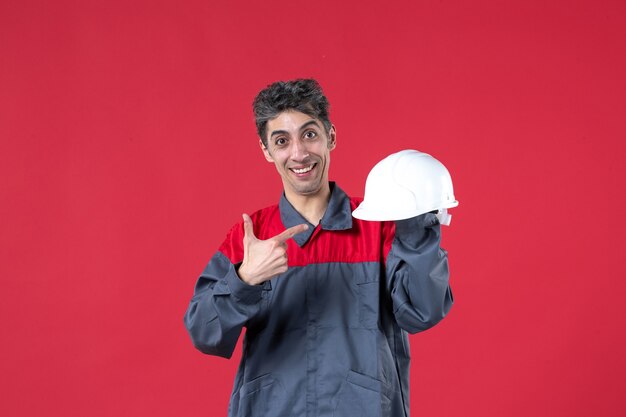 Front view of smiling young worker in uniform and pointing hard hat on isolated red wall