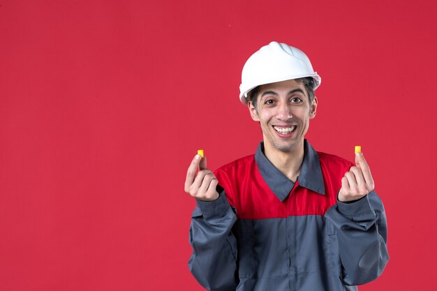 Вид спереди улыбающегося молодого работника в униформе с каской и затычками для ушей на изолированной красной стене
