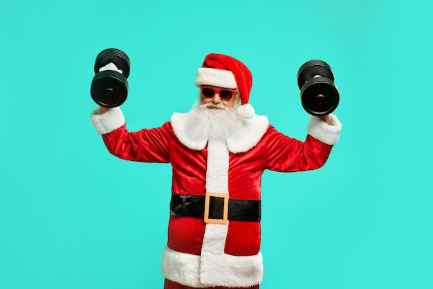 Front view of sportive Santa Claus holding dumbbels. Isolated portrait of funny senior man in christmas costume and sunglasses posing 