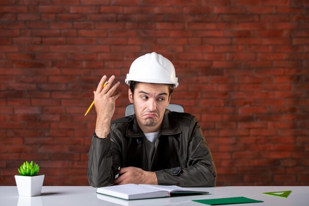 Front view stressed male engineer sitting behind his working place and writing notes document plan job business corporate property builder agenda