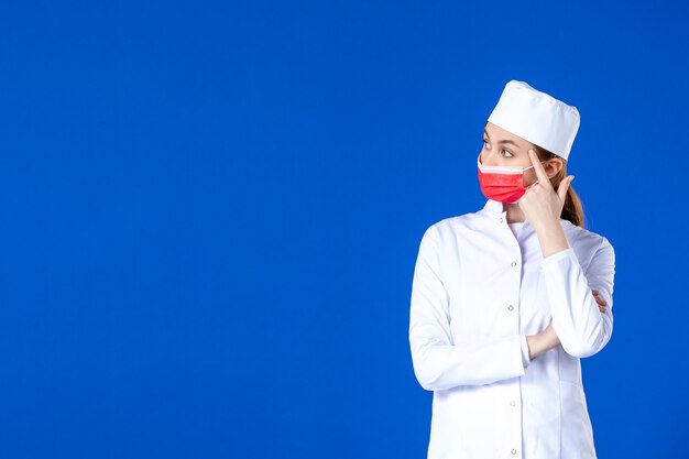 Front view stressed young nurse in medical suit with red protective mask on blue wall
