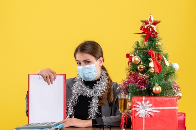 Front view strong girl with medical mask sitting at the table xmas tree and gifts cocktail