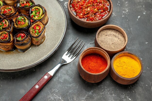 Front view stuffed aubergine rolls in white oval plate fork oil adjika in small bowl different spices on grey