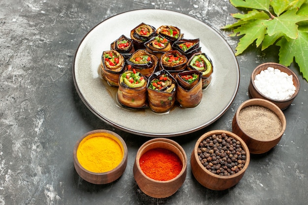 Free photo front view stuffed eggplant rolls on white oval plate different spices in small bowls leaves on grey background