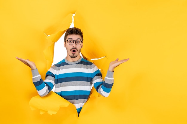 Front view stunned young man peeping through hole in paper yellow wall