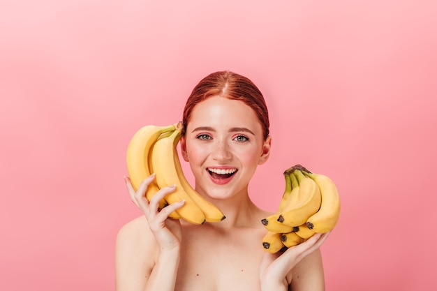 Front view of stunning ginger girl with bananas. Studio shot of happy nude woman holding tropical fruits on pink background.