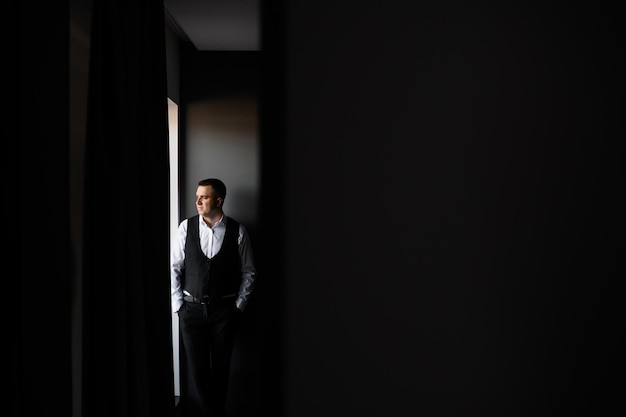 Front view of stylish bridegroom in wedding outfits standing near window keeping hands in pocket and looking away during wedding morning in cozy and dark room