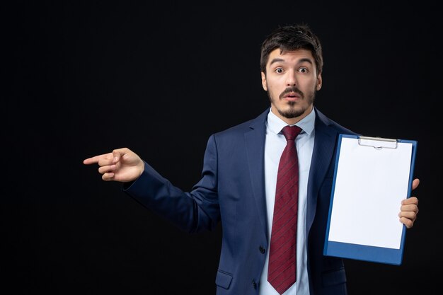 Front view of surprised adult in suit holding free space for writing and pointing something on the right side on isolated dark wall