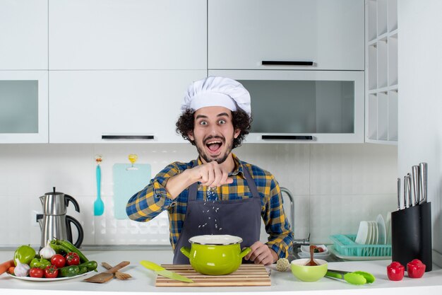 Front view of surprised chef with fresh vegetables adding salt into pot in the white kitchen