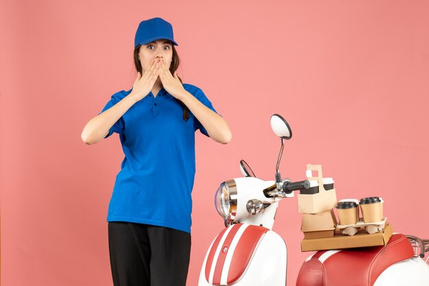 Front view of surprised courier lady standing next to motorcycle with coffee and small cakes on it on pastel peach color background