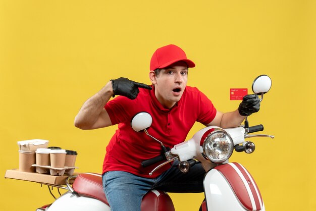 Front view of surprised courier man wearing red blouse and hat gloves in medical mask delivering order sitting on scooter pointing bank card