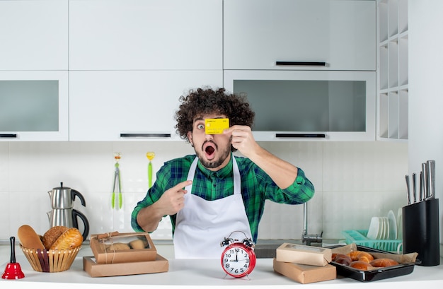 Free photo front view of surprised crazy emotional funny young man standing behind the table various pastries on it and showing bank card in the white kitchen