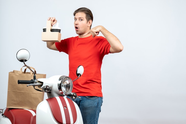 Front view of surprised delivery man in red uniform standing near scooter pointing order on white background