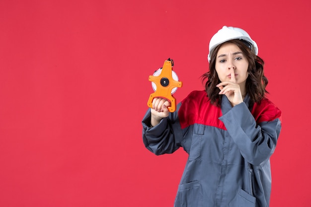 Front view of surprised female architect in uniform with hard hat holding measuring tape and making silence gesture on isolated red background