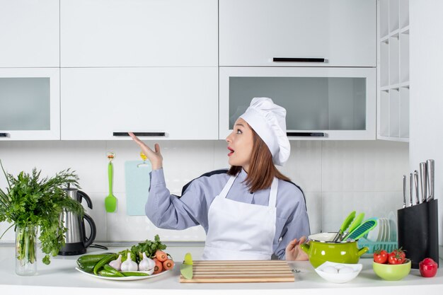 Front view of surprised female chef and fresh vegetables pointing something on the right side in the white kitchen