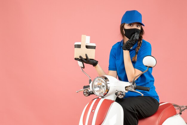 Вид спереди удивленного доставщика женского пола в медицинской маске и перчатках, сидящего на скутере, доставляющего заказы на пастельно-персиковом фоне