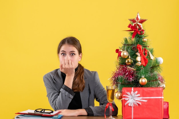 机に座っている正面図驚いた女の子は彼女の口に手を置いたクリスマスツリーとギフトカクテル