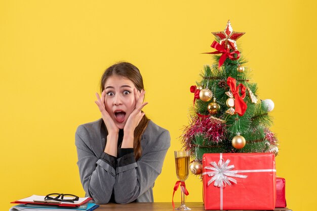 크리스마스 트리와 선물 칵테일 근처에 그녀의 뺨에 손을 댔을 책상에 앉아 전면보기 놀란 소녀