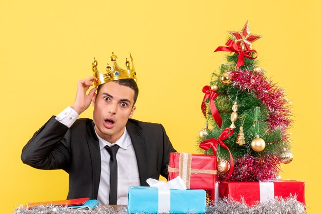 Front view surprised handsome man with crown sitting at the table xmas tree and gifts