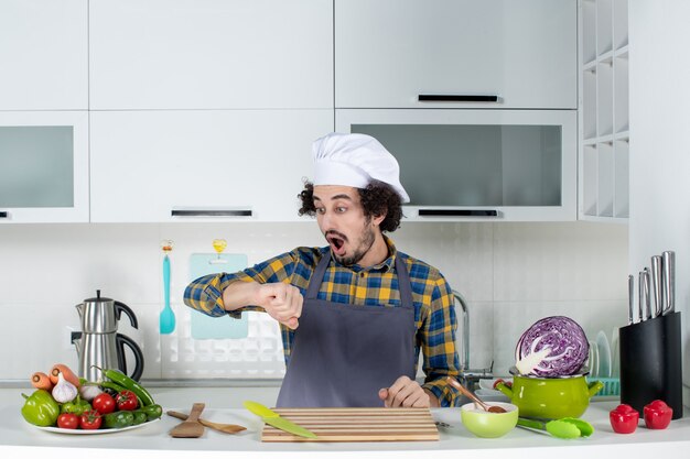 新鮮な野菜とキッチンツールで調理し、白いキッチンで彼の時間をチェックして驚いた男性シェフの正面図