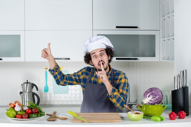 新鮮な野菜とキッチンツールで調理し、白いキッチンで沈黙とOKジェスチャーを作る驚いた男性シェフの正面図