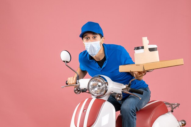 Front view of surprised male delivery person in mask wearing hat sitting on scooter delivering orders on pastel peach background