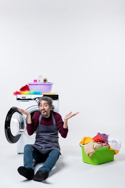 Front view surprised male housekeeper sitting in front of washer laundry basket on white wall