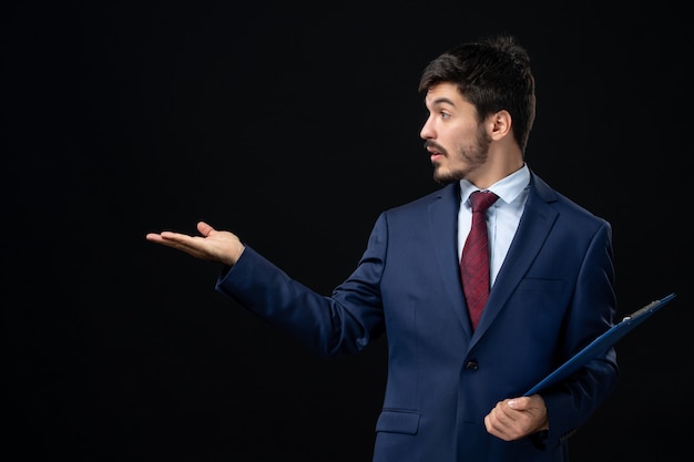 Free photo front view of surprised male office worker in suit holding documents and pointing something on the right side on isolated dark wall