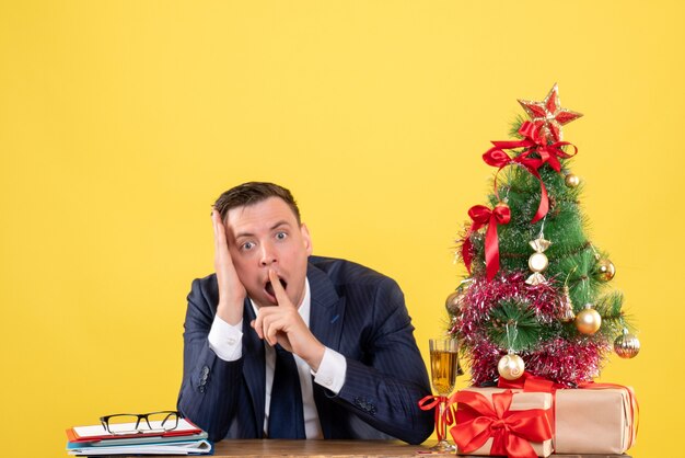 Front view of surprised man sitting at the table near xmas tree and presents on yellow