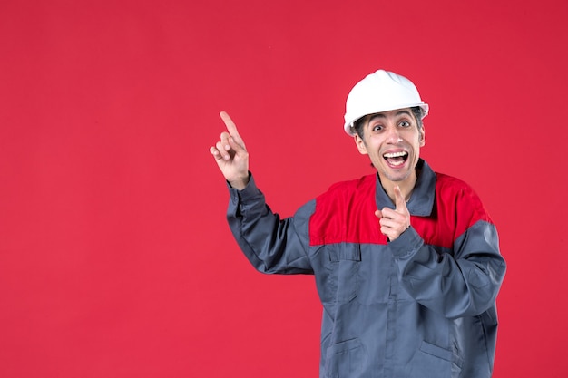 Front view of surprised smiling young worker in uniform with hard hat pointing up on isolated red wall